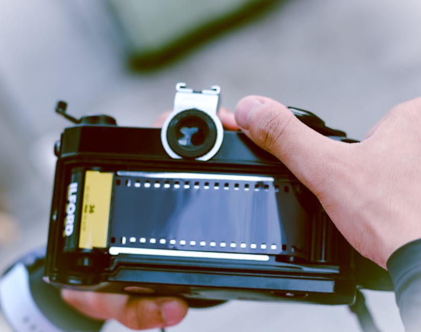 A close-up image showing a vintage film camera positioned on top of numerous rolls of photographic film. The camera is black with a textured surface, and the back of the camera faces the viewer, revea