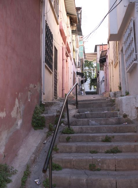 A narrow outdoor staircase made of concrete steps with visible wear and some patches of grass and dirt between them, ascending between old, weathered residential buildings with pastel-colored facades.