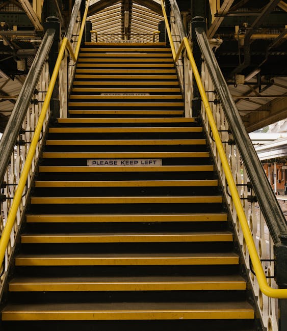 A set of metal stairs with black treads and yellow safety edges leading up to a pedestrian walkway within a railway station structure. The stairs are flanked by yellow handrails on both sides. Several