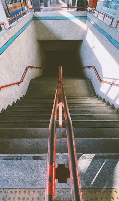 A set of metal stairs with black treads and yellow safety edges leading up to a pedestrian walkway within a railway station structure. The stairs are flanked by yellow handrails on both sides. Several