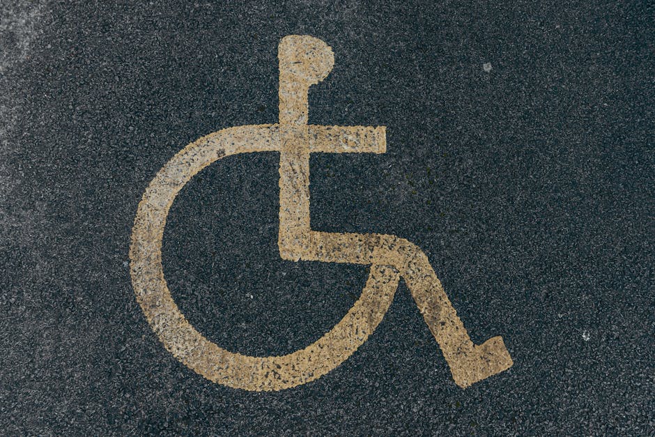 A close-up view of a designated disabled parking space on asphalt pavement, featuring a yellow wheelchair symbol painted onto the surface. The parking spot is adjacent to a curb, with part of a stripe