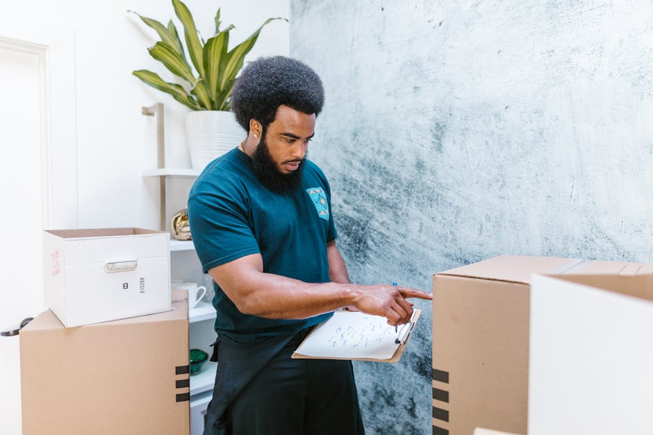 A young professional man wearing a navy blue work jumpsuit and a black bandana with white patterns is inside a well-lit room with a textured light blue wall. He is holding a clipboard and pen, appeari