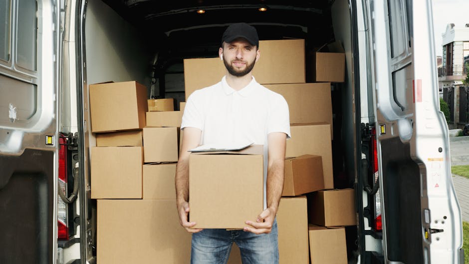 Two men are engaged in a home relocation task outdoors on a brick driveway, preparing to load a large cardboard box into the back of a white moving van parked next to a residential property with a bri